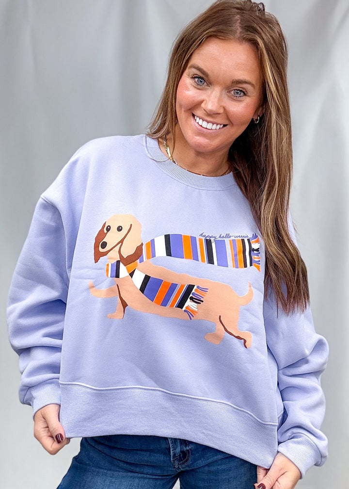 Woman wearing periwinkle sweatshirt with doxie graphic with scarf on front with jeans on plain background