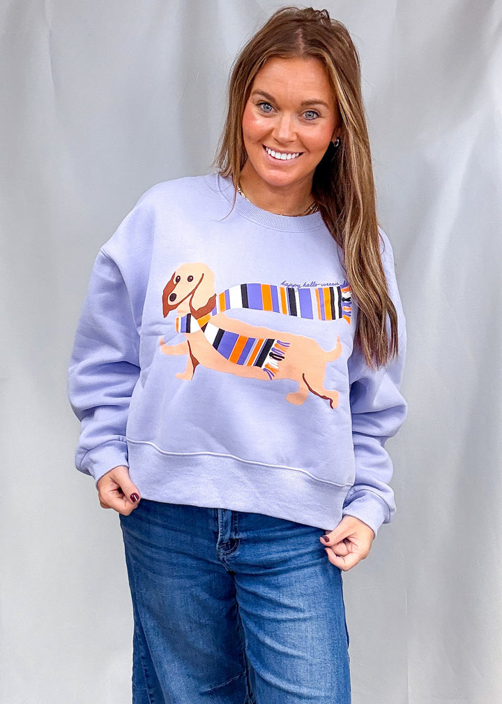A woman wearing a light lavender crewneck sweatshirt with a cartoon dachshund graphic wearing a colorful striped scarf, paired with wide-leg blue jeans, standing against a white draped fabric backdrop.
