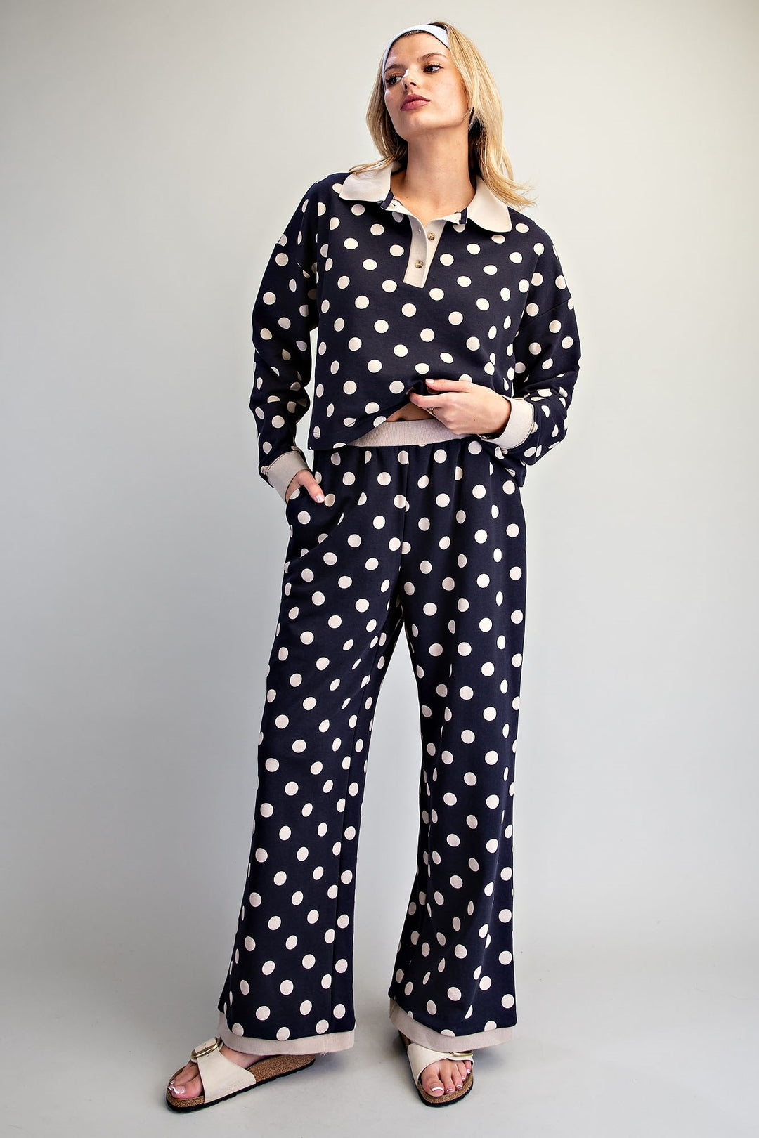 A woman modeling a matching two-piece sleepwear set featuring a navy blue and white polka dot pattern with light beige trim, consisting of a long-sleeve collared top and matching wide-leg pants, set against a plain light gray studio background.