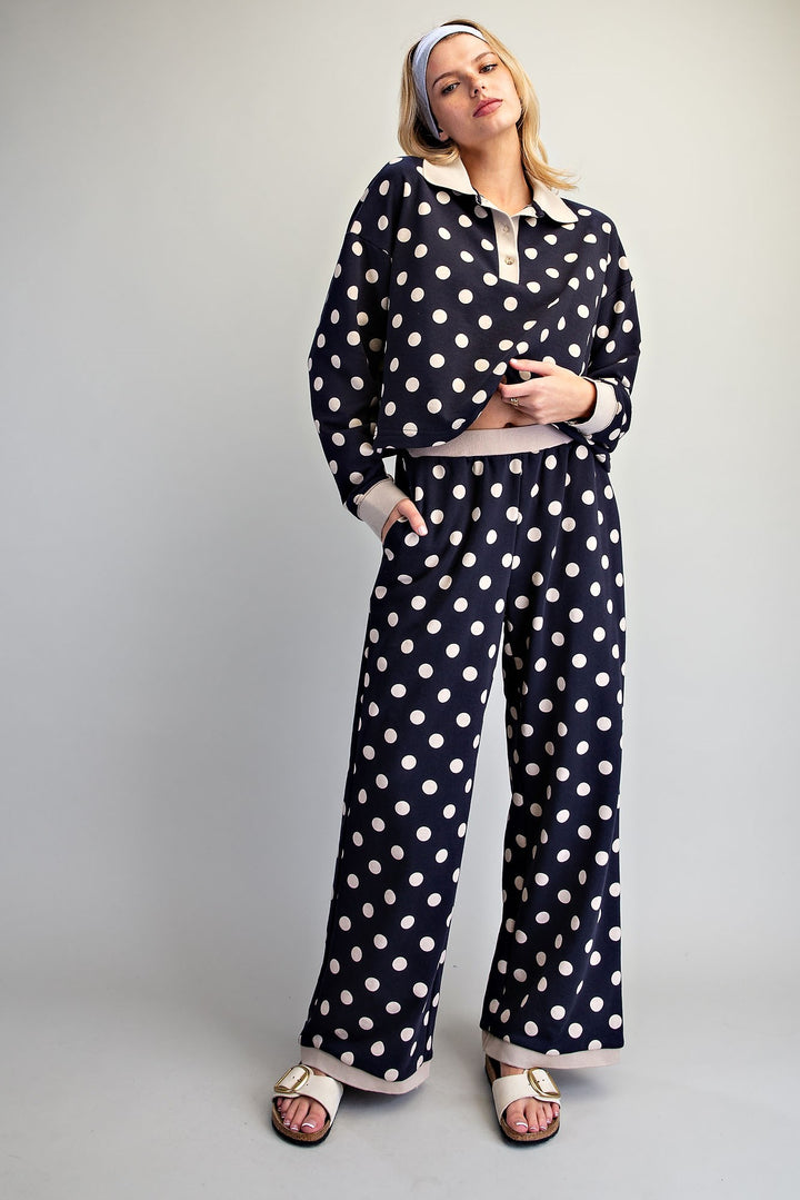 A woman modeling a matching two-piece sleepwear set with a navy blue and white polka dot pattern, featuring a long-sleeve collared top and wide-leg pants, styled with a matching headband and beige slide sandals against a neutral studio background.