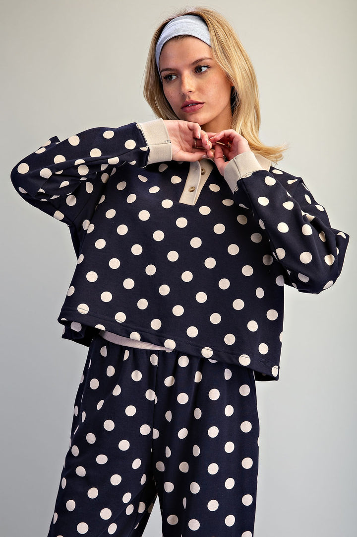 A woman modeling a matching navy blue and white polka-dot pajama set, featuring a cropped long-sleeve henley top with a button placket and matching pants, against a plain light gray background.