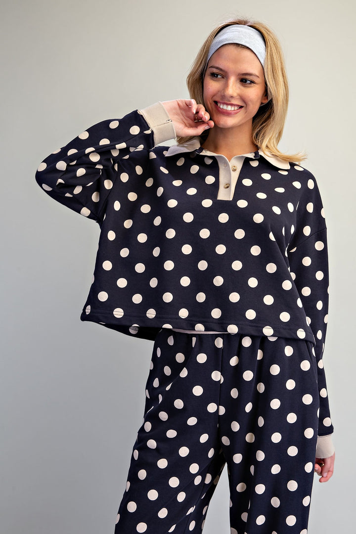 A woman modeling a matching two-piece pajama set with a navy blue base and large white polka dots. The set includes a long-sleeve collared top with a button placket and matching drawstring pants.