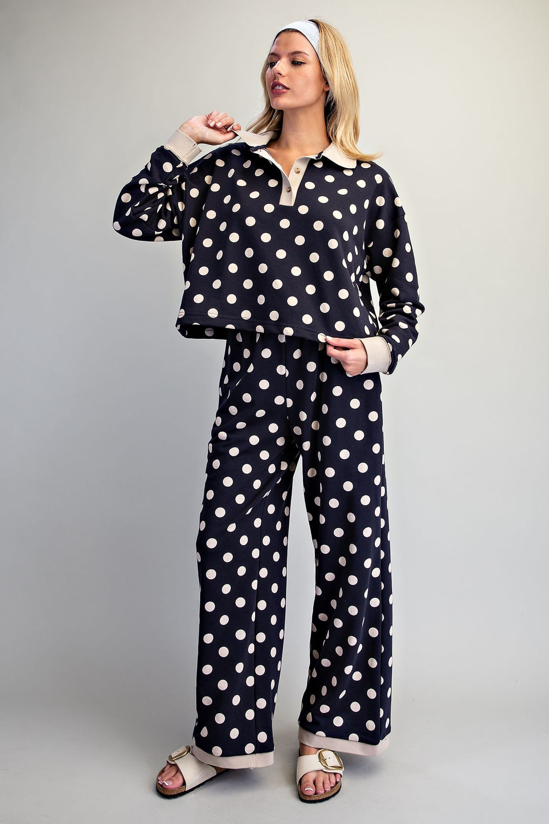 A woman modeling a matching two-piece pajama set with a navy blue and white polka dot pattern, featuring a long-sleeve collared top and wide-leg pants, styled with white slide sandals and a white headband against a neutral gray background.