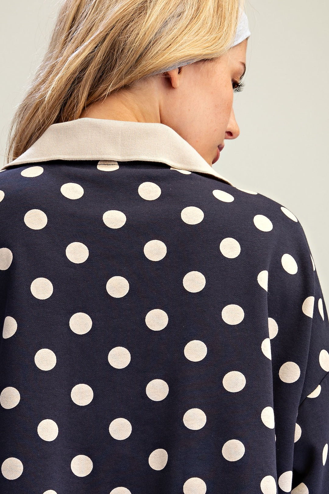 Close-up back view of a woman wearing a navy blue top with large white polka dots and a contrasting light beige collar, against a plain light gray background.