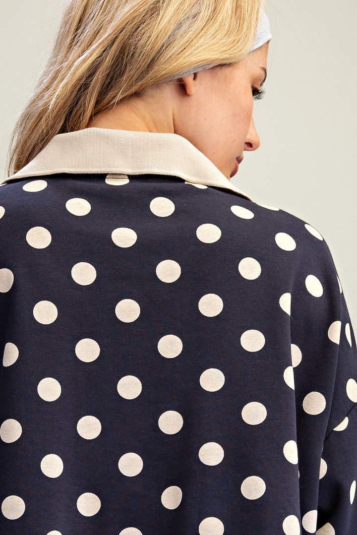 Close-up back view of a woman wearing a navy blue top with large white polka dots and a contrasting light beige collar, against a plain light gray background.