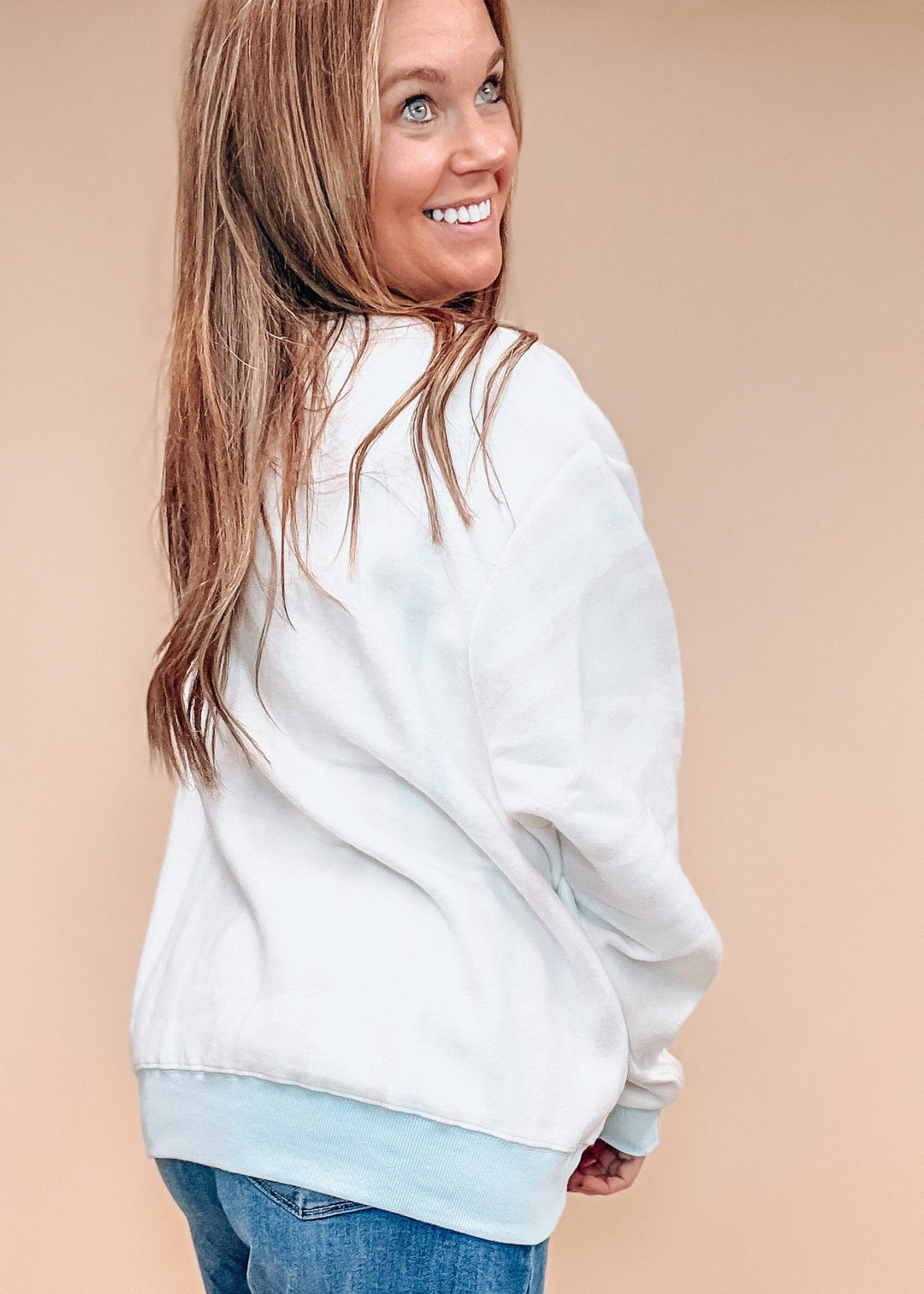 A woman wearing a white crewneck sweatshirt with a light blue ribbed hem, paired with blue jeans, shown from a side-back angle against a solid beige background.
