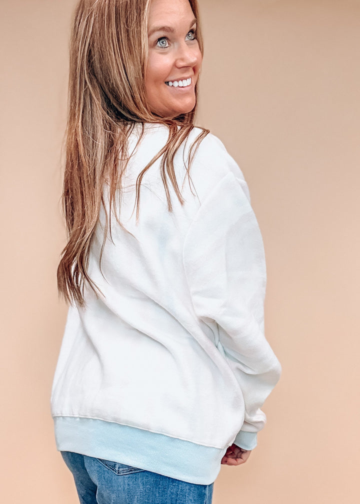 A woman wearing a white crewneck sweatshirt with a light blue ribbed hem, paired with blue jeans, shown from a side-back angle against a solid beige background.
