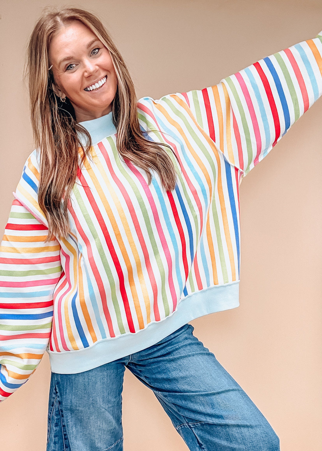 A woman wearing a colorful vertically striped long-sleeve sweatshirt with blue jeans, posing against a solid beige background.