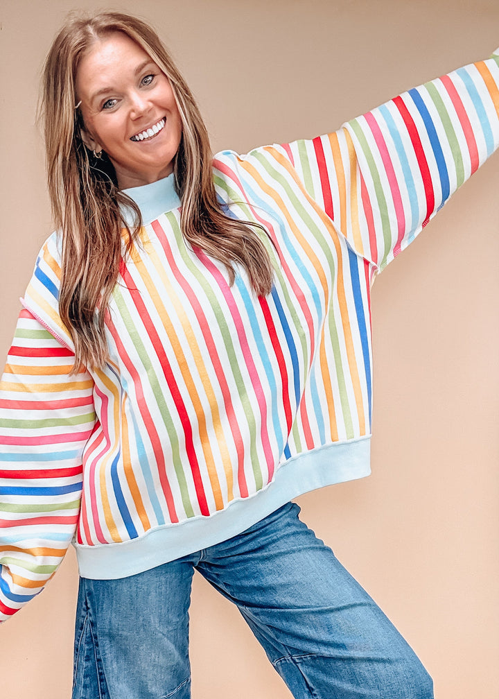 A woman wearing a colorful vertically striped long-sleeve sweatshirt with blue jeans, posing against a solid beige background.