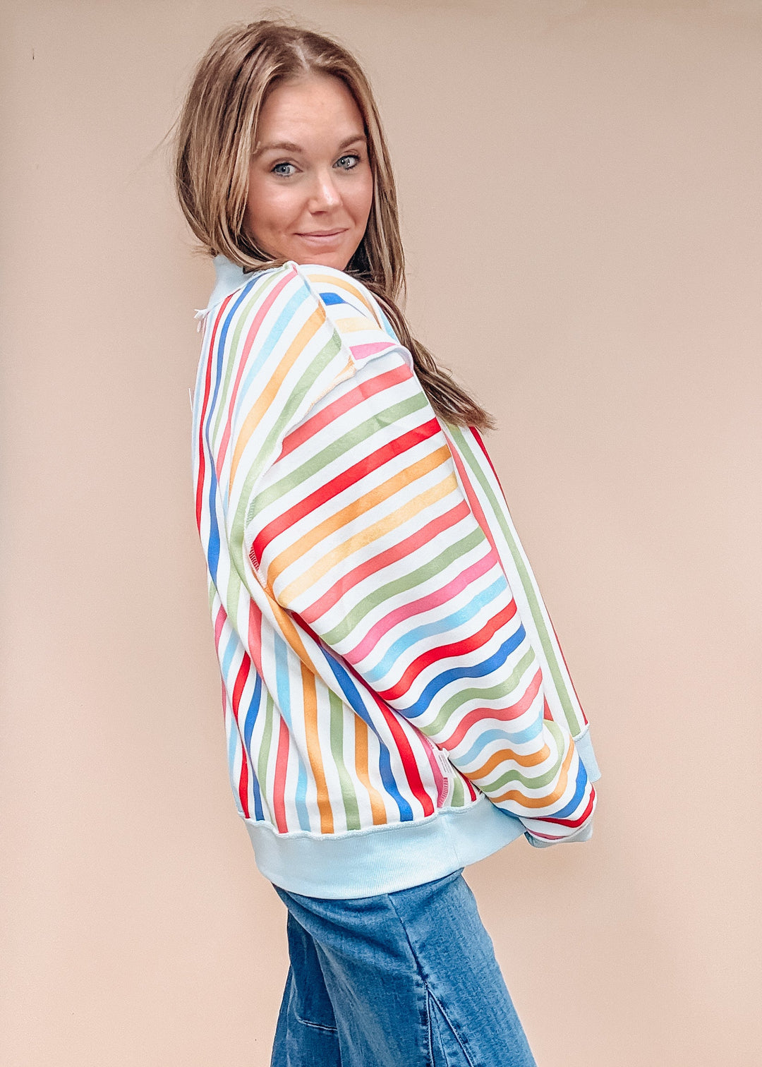 A woman wearing a colorful striped cropped sweatshirt and a blue denim skirt, posing in a studio with a beige background.