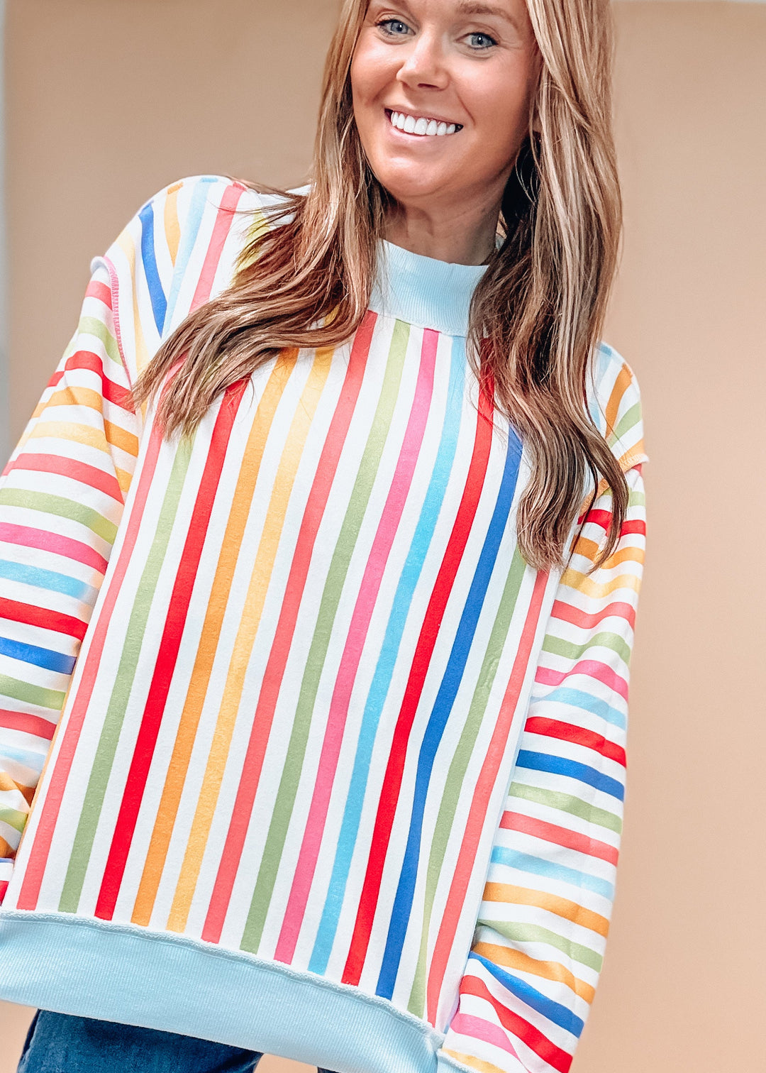 A woman wearing a colorful striped long-sleeve sweatshirt with a light blue mock neck and hem, paired with blue jeans, against a solid beige background.