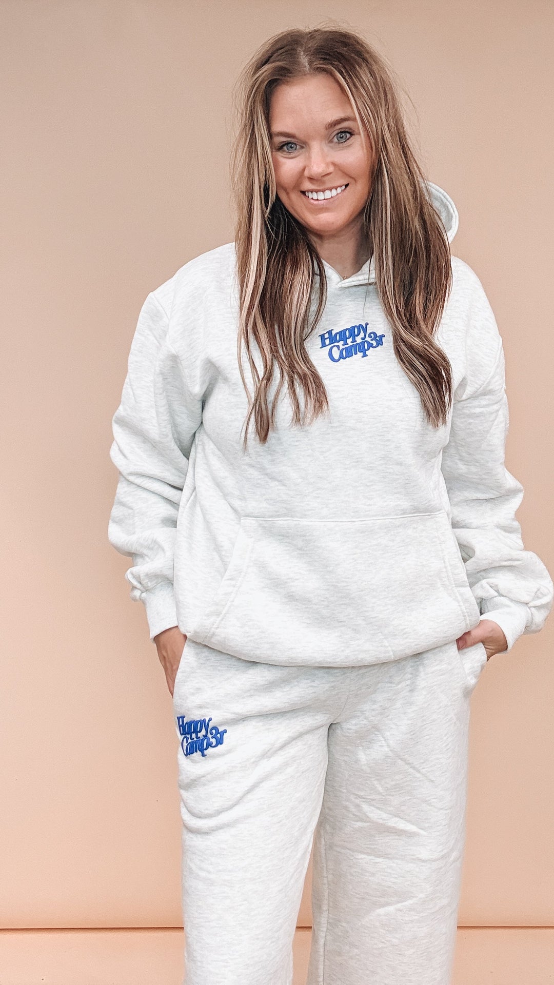 A woman modeling a matching light heather gray sweatshirt and jogger pants set with blue 'Happy Camp3r' text graphics on the chest and thigh, posed against a solid peach studio backdrop.