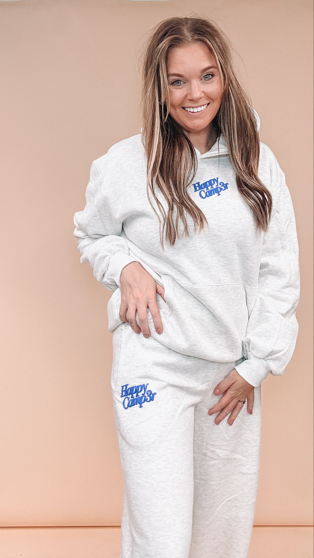 A woman modeling a matching light gray heather sweatshirt and jogger sweatpants set with blue 'Happy Camp3r' text printed on the chest and thigh, set against a solid peach studio backdrop.