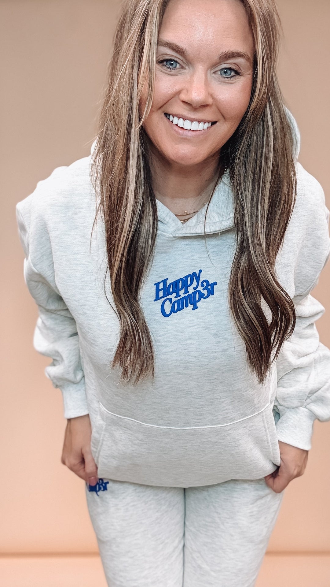 A smiling woman with long light brown hair wears a light heather gray hoodie and matching sweatpants set, both featuring a blue &