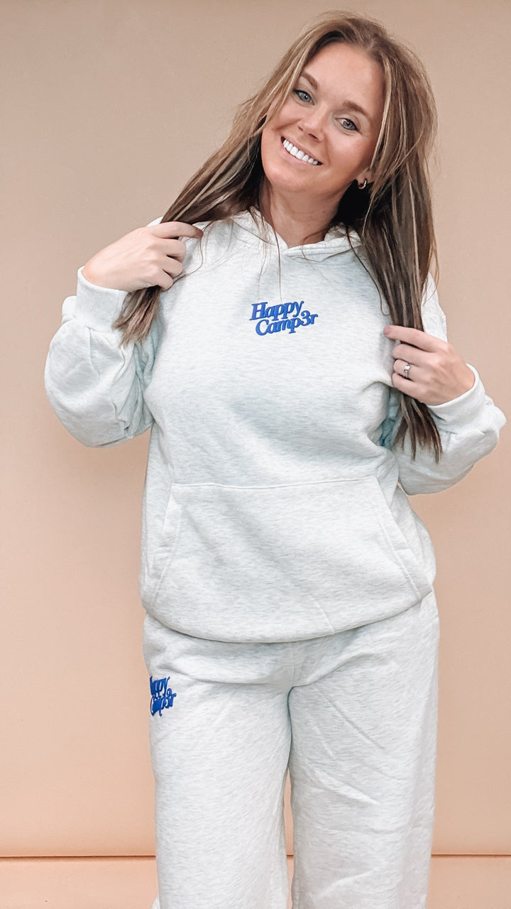 A woman modeling a light heather gray matching lounge set consisting of a hoodie and sweatpants, both featuring a blue embroidered &