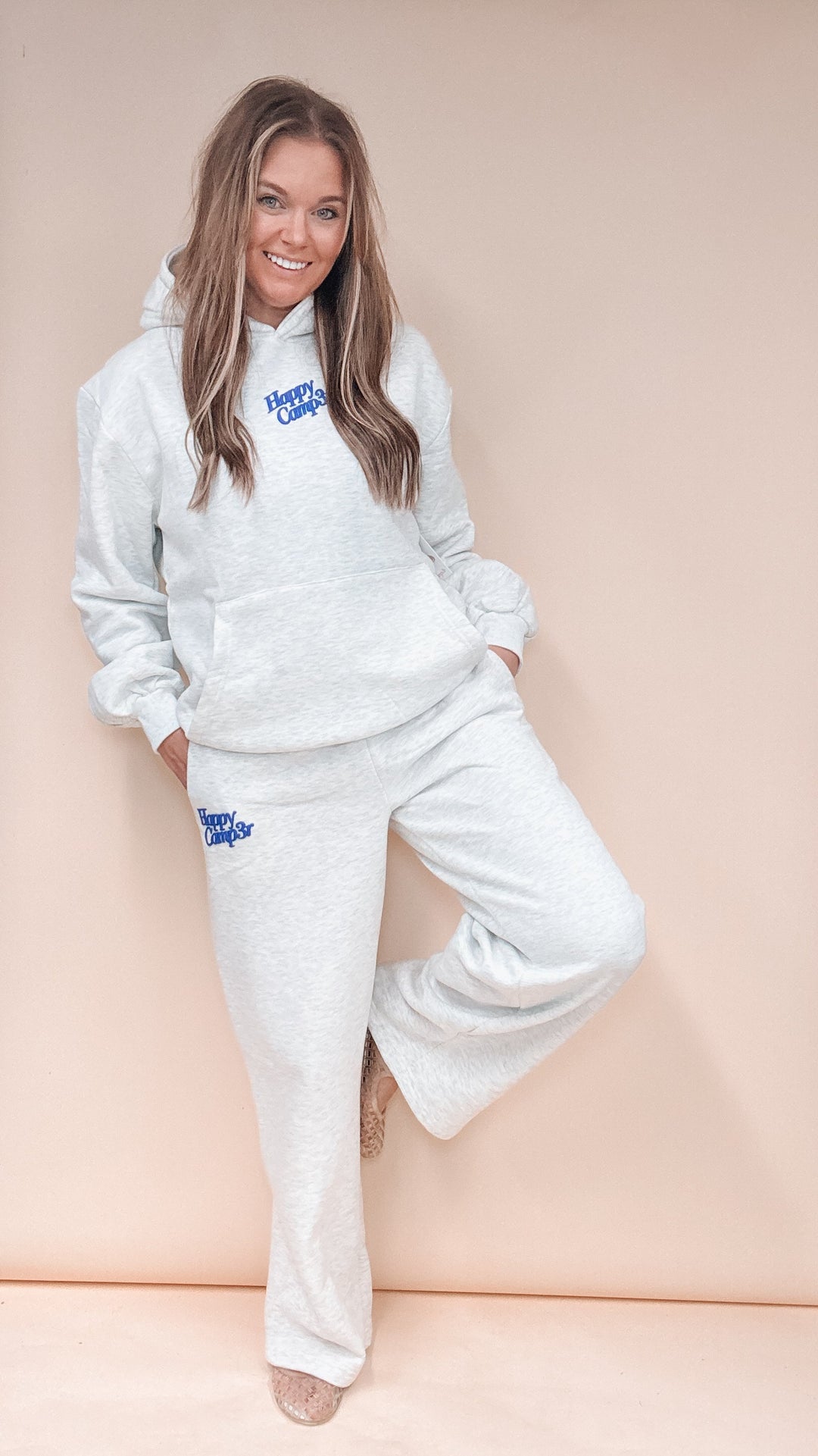 A woman modeling a light heather gray two-piece loungewear set with blue 'Happy Camp3r' text on the chest and thigh, posing against a solid pale peach studio backdrop.