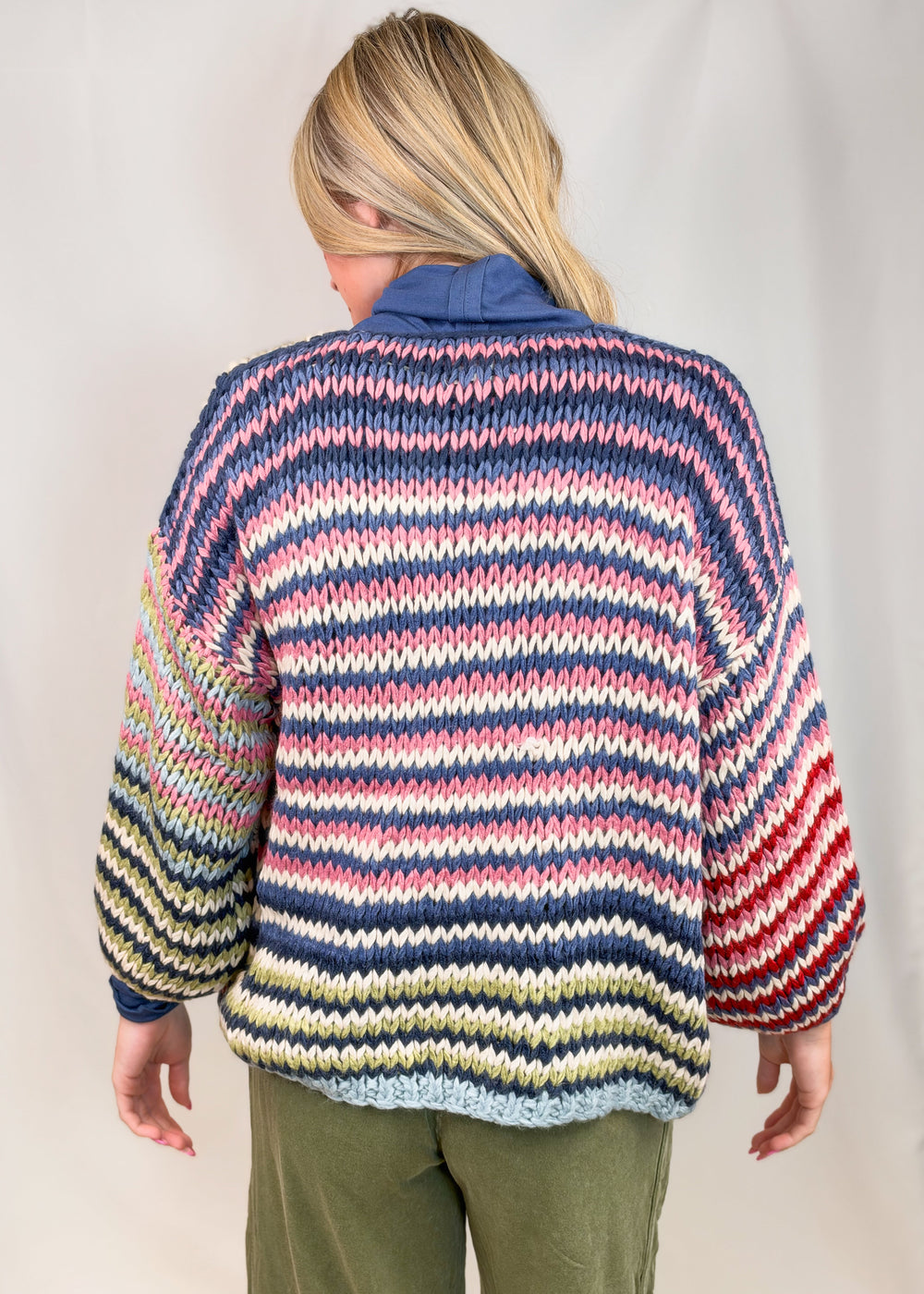 Woman shown from back wearing colorful striped cardigan with green pants on plain background