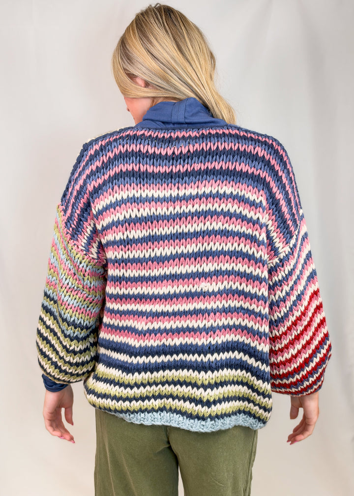 Woman shown from back wearing colorful striped cardigan with green pants on plain background