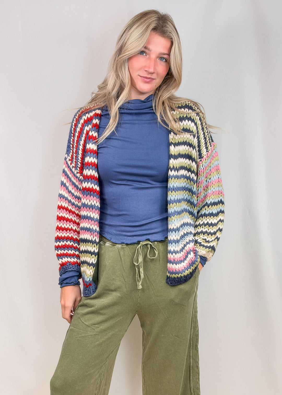 Woman wearing colorful stripe cardigan sweater with blue shirt and green pants on plain background