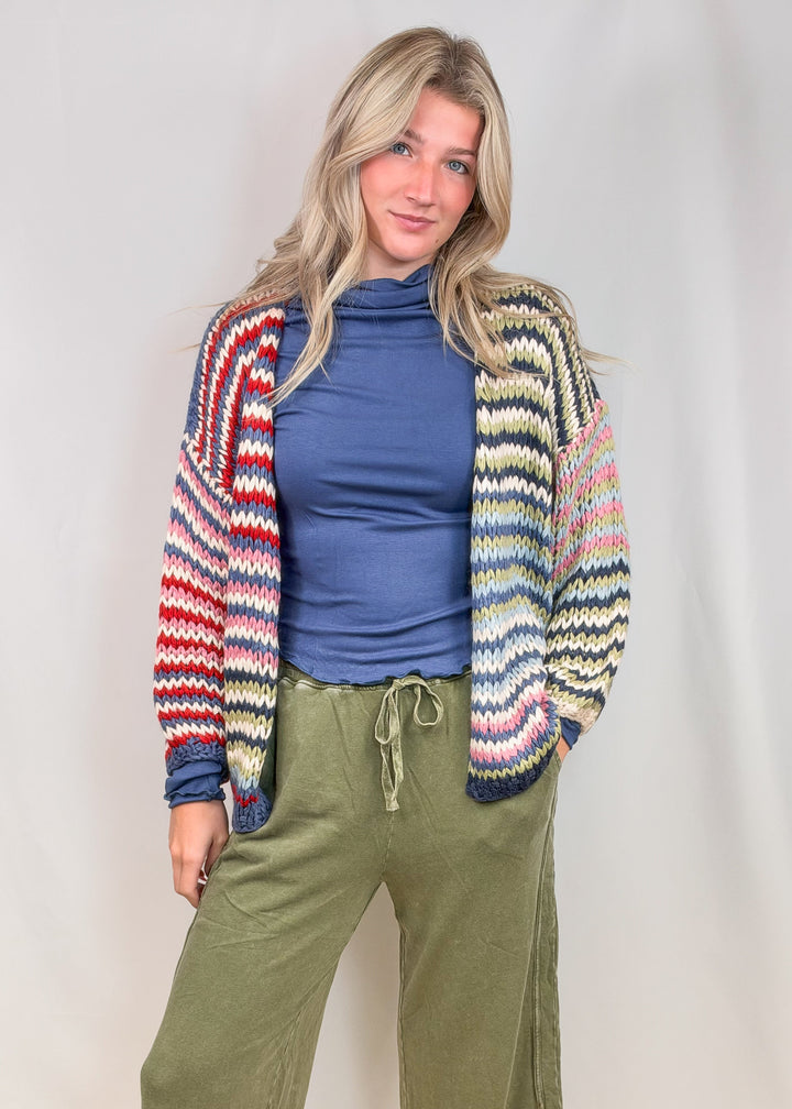 Woman wearing colorful stripe cardigan sweater with blue shirt and green pants on plain background