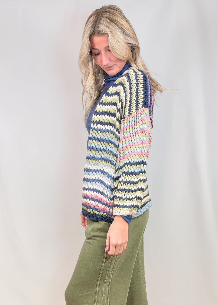 Woman shown from side view wearing colorful stripe cardigan sweater with green pants on plain background
