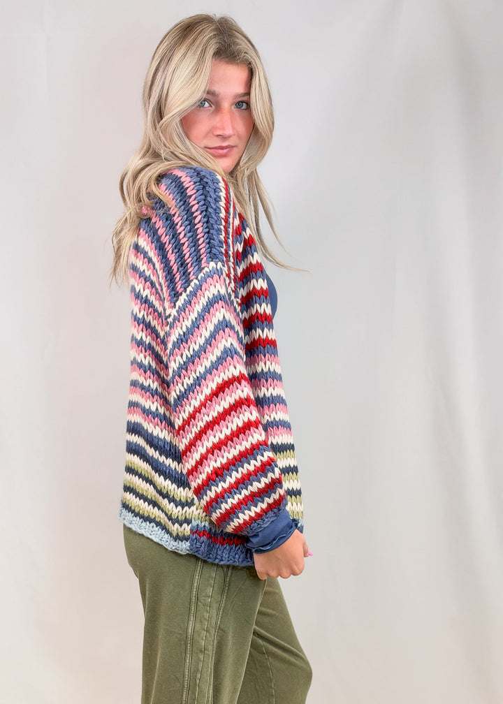 Woman shown from side view wearing colorful stripe cardigan with green pants on plain background