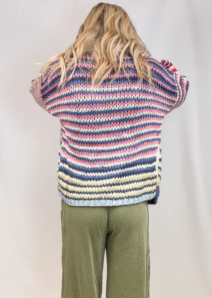 Woman shown from back view wearing colorful stripe cardigan and green pants on plain background