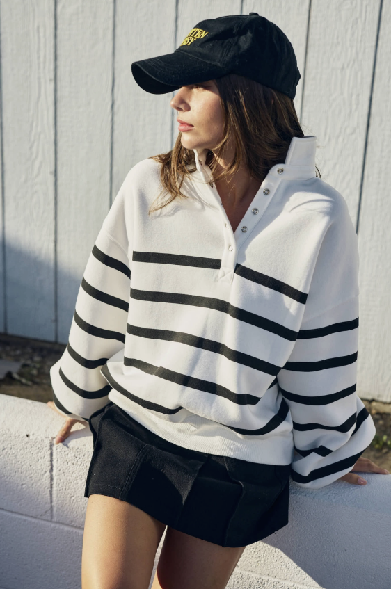 A woman wearing a white long-sleeve rugby-style pullover with bold black horizontal stripes, a black baseball cap, and a black skirt, leaning against a white wall in front of a white paneled wall.