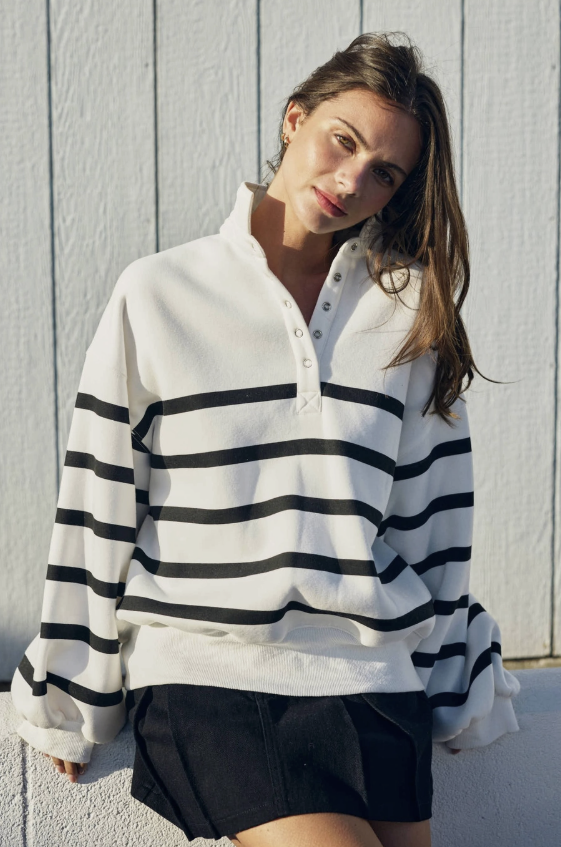 A woman wearing a white long-sleeve rugby-style pullover with black horizontal stripes, paired with black shorts, leaning against a white paneled wall.