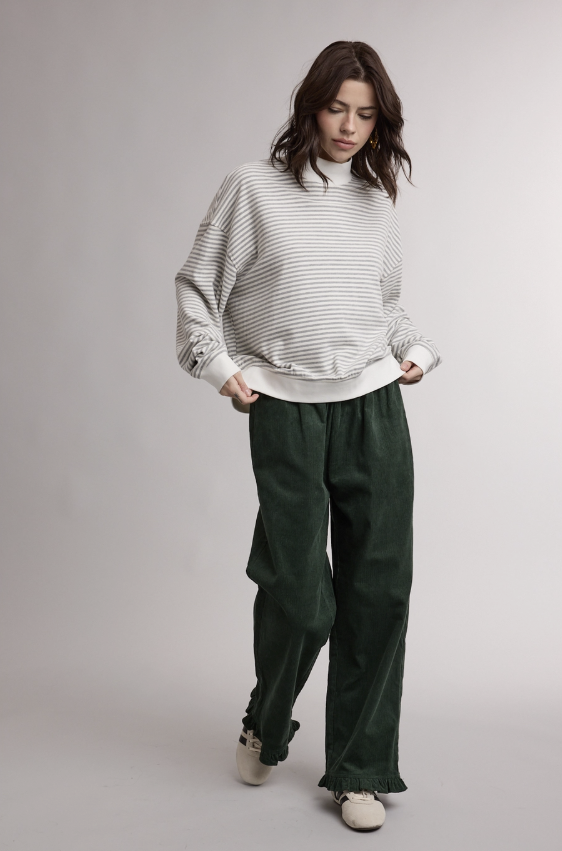 A woman modeling a white and gray striped long-sleeve sweatshirt with distressed details on the cuffs, paired with dark green wide-leg trousers and white sneakers, against a light gray studio background.