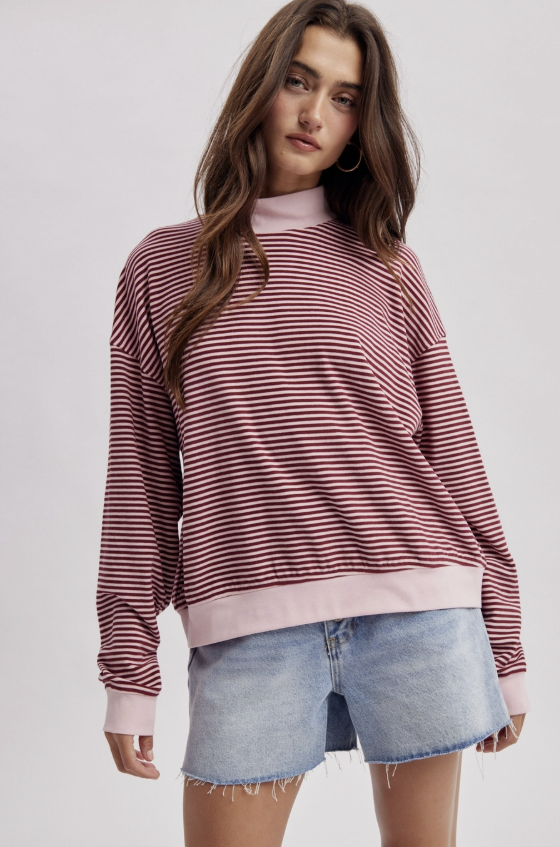 A woman modeling a long-sleeve crewneck sweatshirt with horizontal pink and white stripes, paired with light-wash denim shorts, against a solid light gray background.