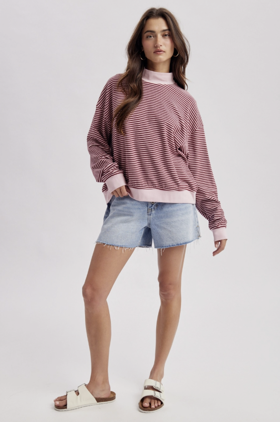 A woman modeling a pink and white striped long-sleeve sweatshirt, paired with light blue denim shorts and white slide sandals, against a light gray studio background.