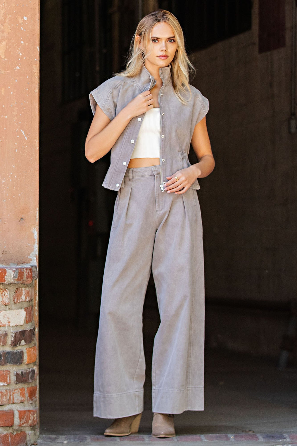 A woman models a light gray linen two-piece set consisting of a sleeveless button-up shirt and matching wide-leg pants, styled with a white crop top and beige heels.