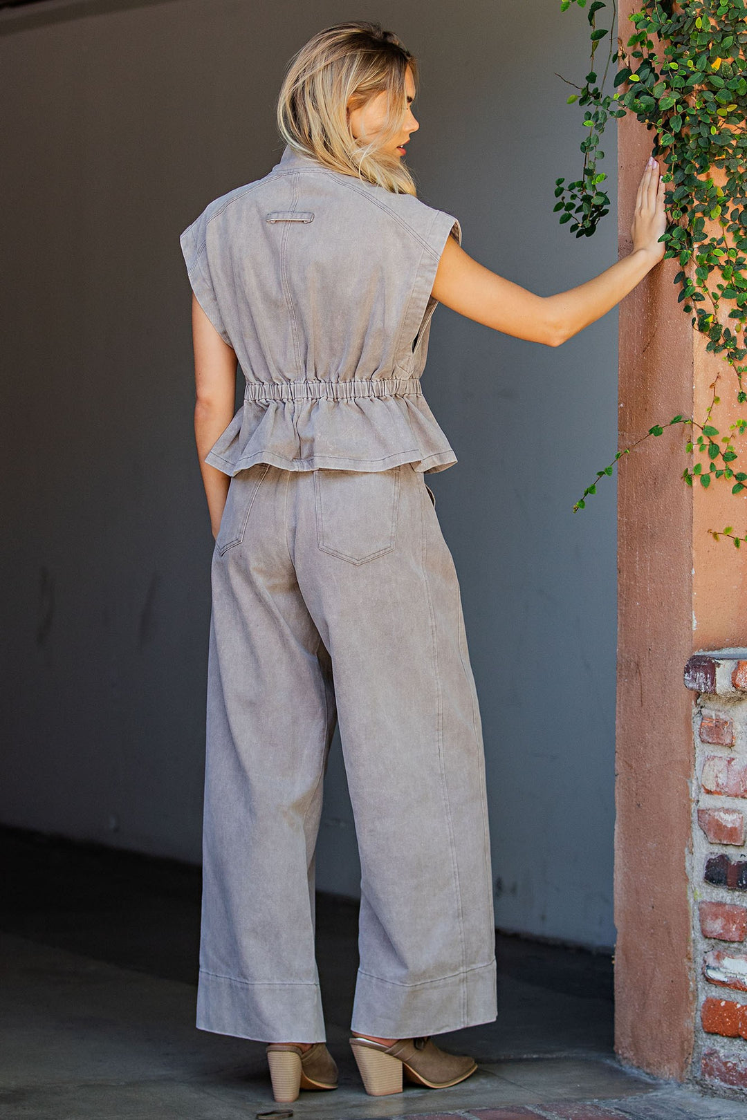 A woman is shown from behind wearing a light gray linen jumpsuit with a ruffled peplum waist and wide-leg pants, standing next to a brick pillar with green ivy.