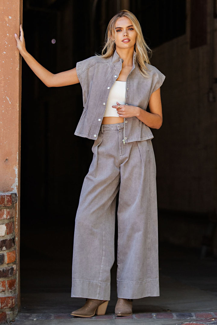 A woman models a light gray linen two-piece set consisting of a sleeveless button-front vest and matching wide-leg pants, styled with a white crop top and brown boots.