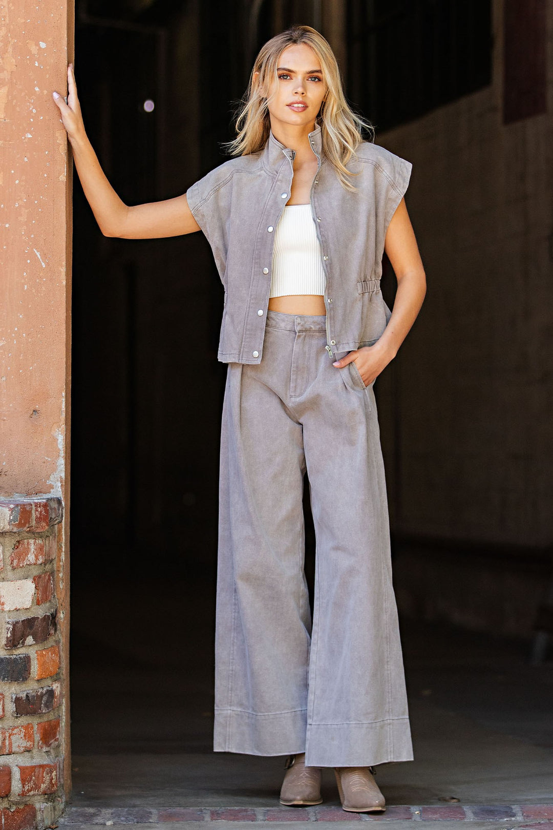 A woman stands in a doorway wearing a light gray sleeveless button-front vest, matching wide-leg pants, a white cropped tank top, and beige flats.