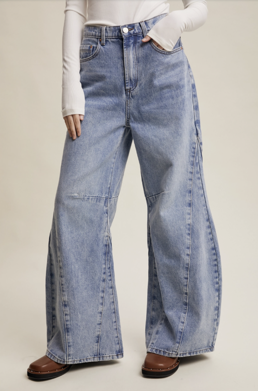 A model wearing light-wash, wide-leg denim jeans with a straight-to-wide leg silhouette, styled with a white long-sleeve top and brown leather shoes against a neutral beige studio background.