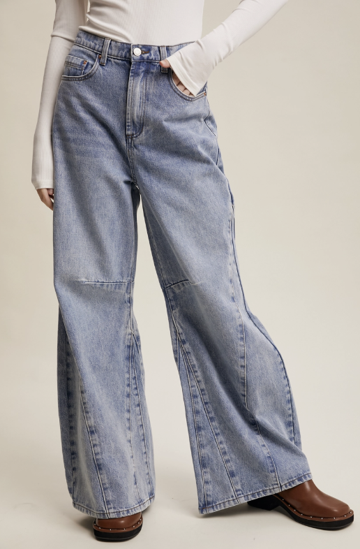 A model wearing light-wash blue wide-leg denim jeans with a raw hem, styled with a white long-sleeve top and brown leather boots against a plain beige studio background.