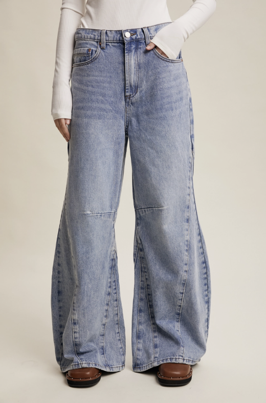 A model wearing light-wash blue wide-leg denim jeans with a straight-to-flared leg and visible seam details, paired with a white long-sleeve top and brown shoes, against a solid beige studio background.