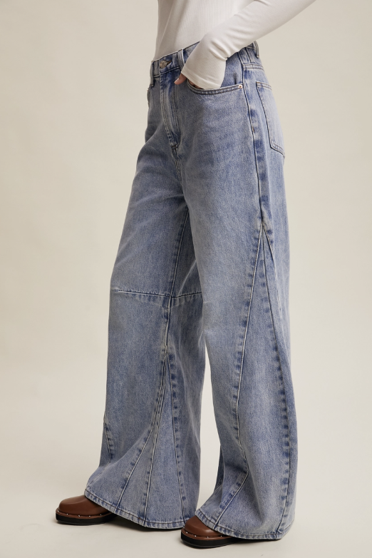 A person wearing light-wash blue wide-leg denim jeans with a white long-sleeve top and brown shoes, shown from the waist down against a plain beige studio background.