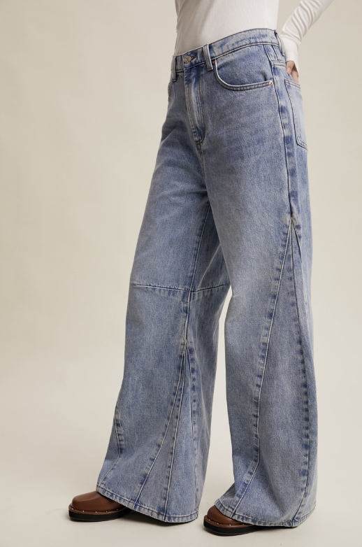 A person wearing light-wash blue wide-leg denim jeans with a white long-sleeve top and brown shoes, shown from the waist down against a plain beige studio background.