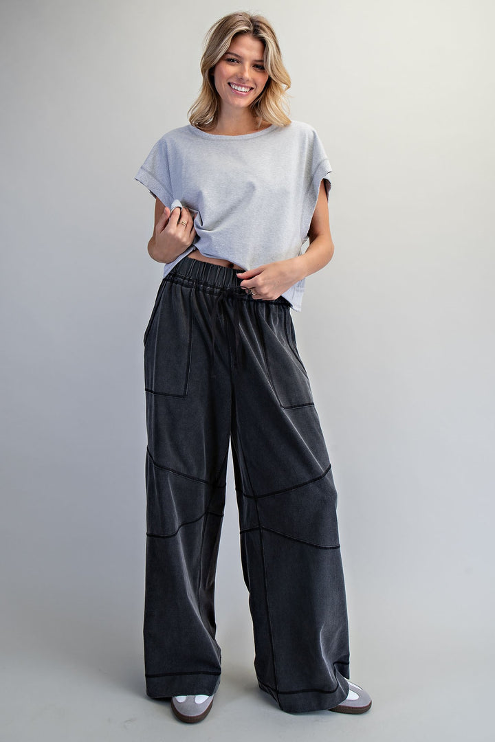 A woman modeling a light gray short-sleeve t-shirt and black wide-leg pants with contrast stitching, standing against a neutral light gray studio background.