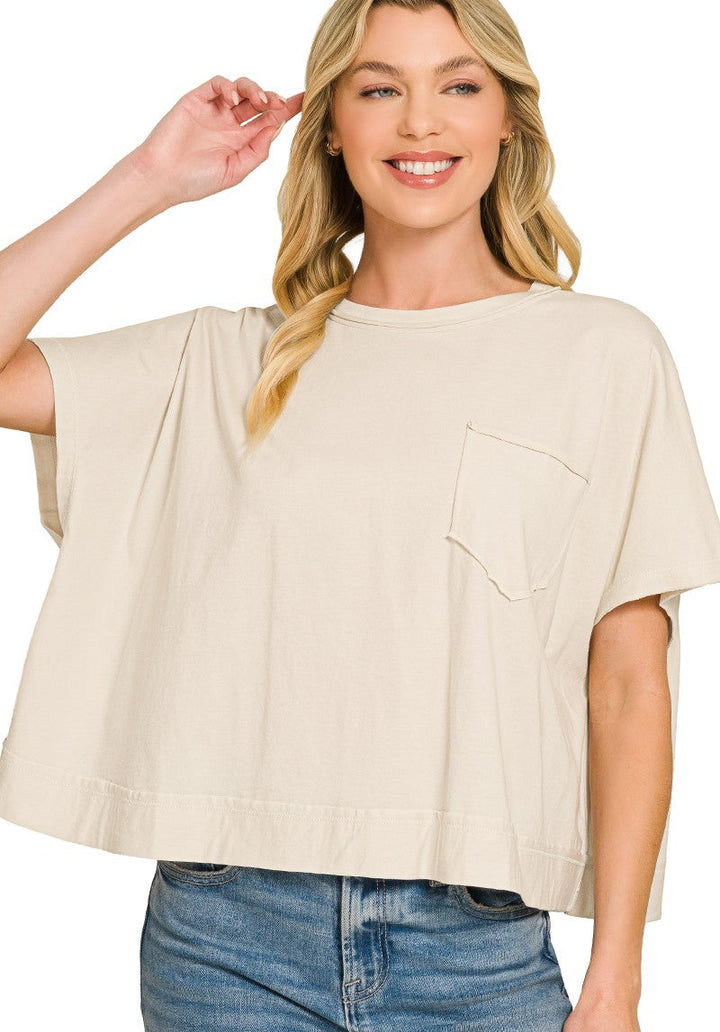 A woman wearing a loose-fitting, short-sleeve sand beige top with a chest pocket, paired with blue jeans, against a plain white background.