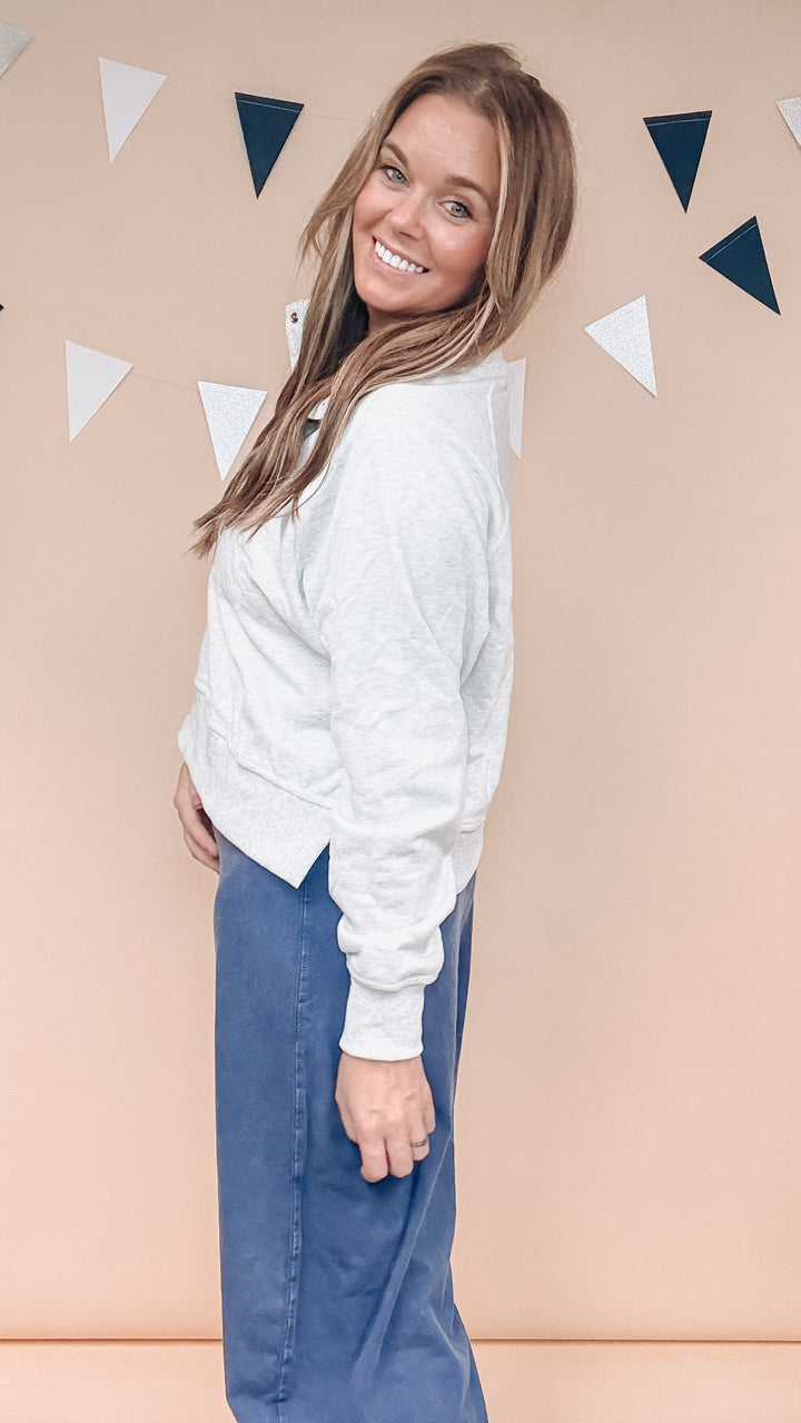 A woman wearing a light gray long-sleeve cropped sweatshirt and blue jeans, posing in front of a peach-colored wall with hanging triangular decorations.
