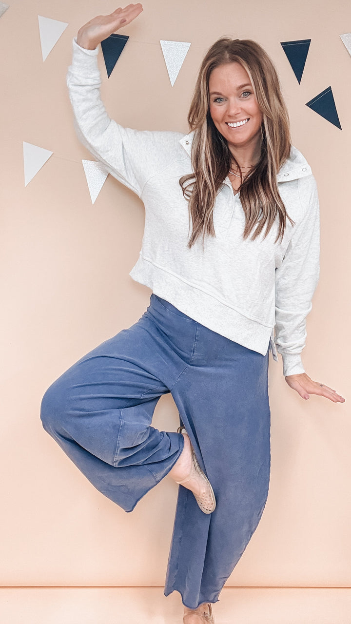 A woman posing in a white cropped sweatshirt and wide-leg blue pants against a peach-colored wall with hanging triangular decorations.