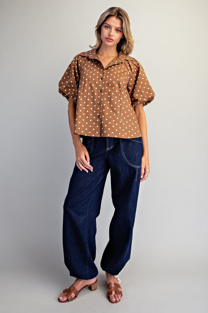 A woman modeling a brown short-sleeve button-up shirt with white polka dots, paired with dark blue wide-leg jeans and brown heeled sandals, against a light gray studio background.