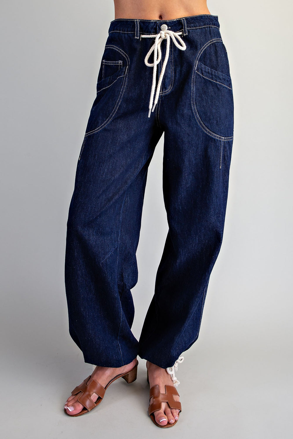 A person wearing dark blue wide-leg denim pants with a white drawstring waist and brown heeled sandals, shown from the waist down against a light gray studio background.