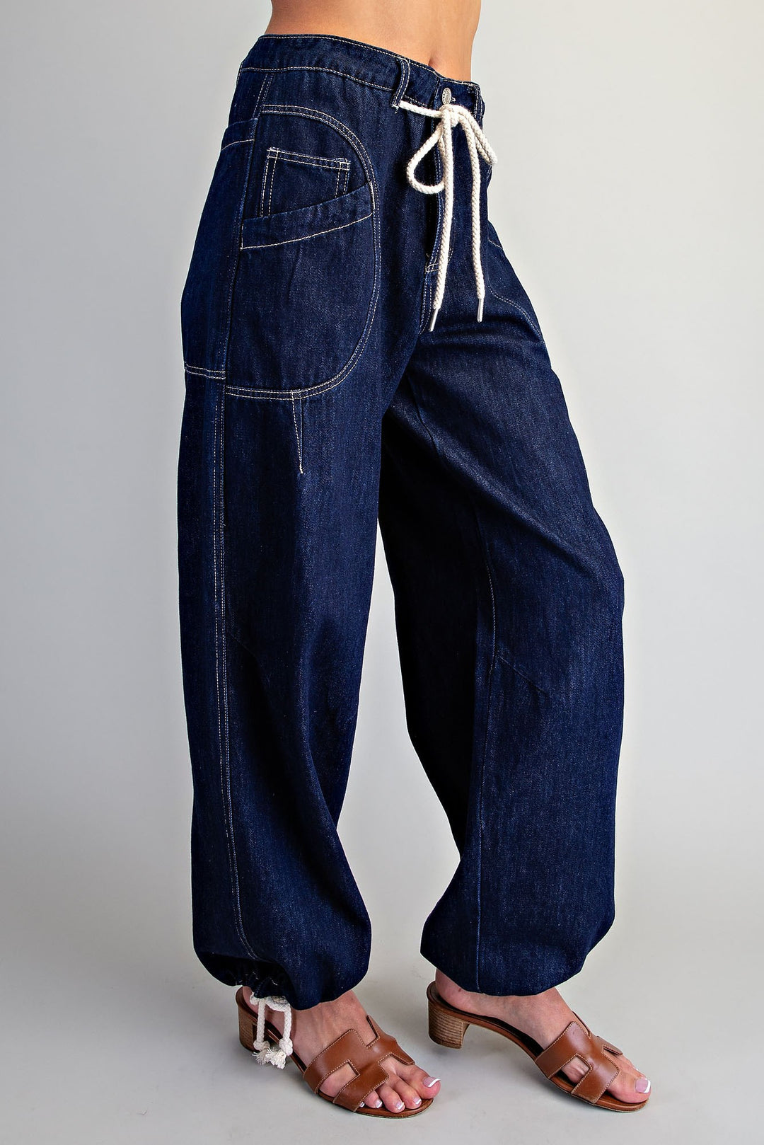 A person wearing dark blue wide-leg denim pants with a white drawstring waist and brown heeled sandals, shown from the waist down against a light gray studio background.