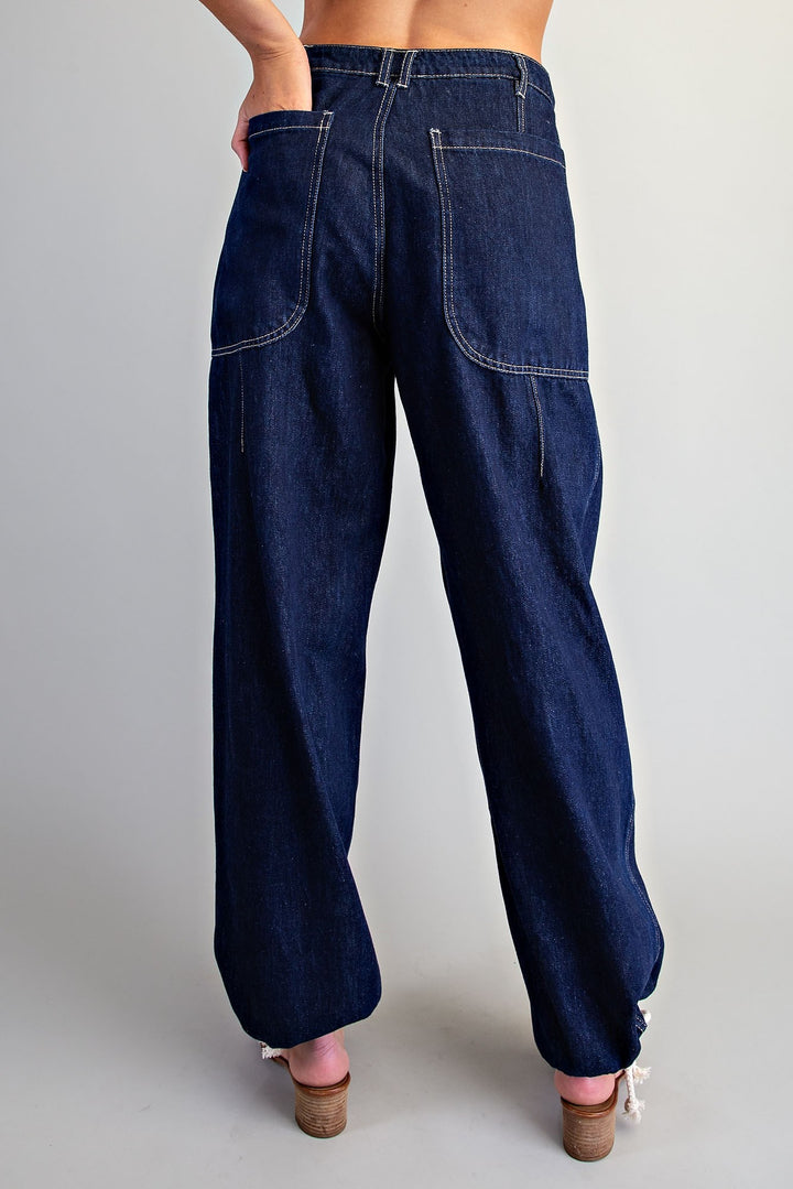 Rear view of a person wearing dark indigo denim jeans with a relaxed, wide-leg fit and cuffed ankles, paired with pink wedge sandals against a light gray studio background.