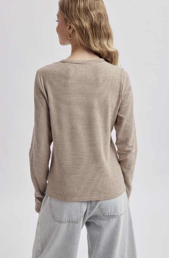 A woman shown from the back wearing a beige long-sleeve top with thin horizontal dark stripes and light-wash jeans, against a plain light gray studio background.